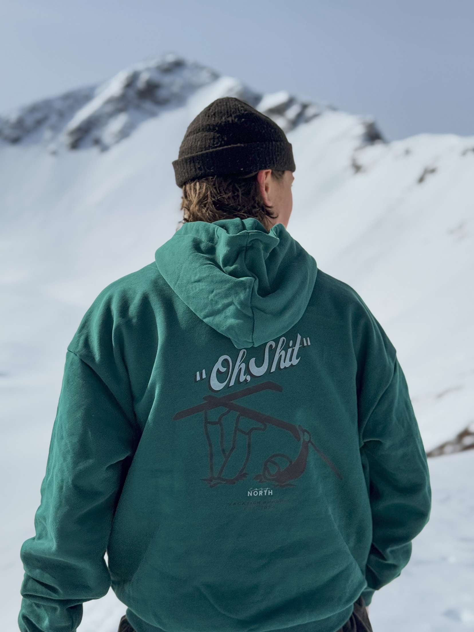 Person i grön CozyNorth hoodie med ”Oh Shit”-tryck på ryggen, fotograferad i snöiga fjäll – streetwear möter skidåkning och après-ski.