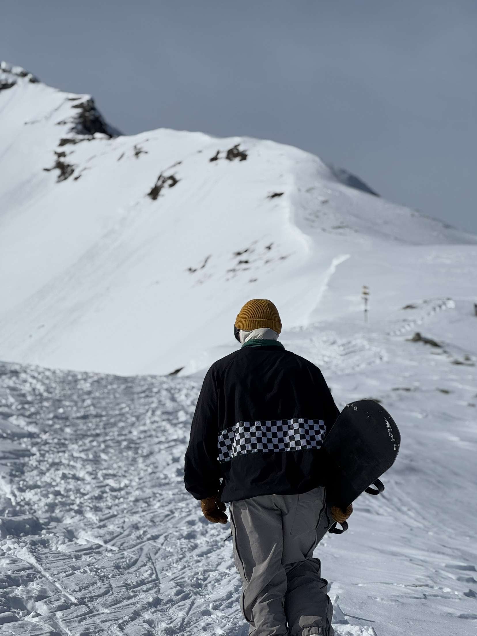 Snowboardåkare på väg uppför snötäckta fjäll med brädan i handen och en cozynorth mössa på sig, en äventyrlig vinterbild som speglar CozyNorths outdoor- och skidåkarlivsstil.