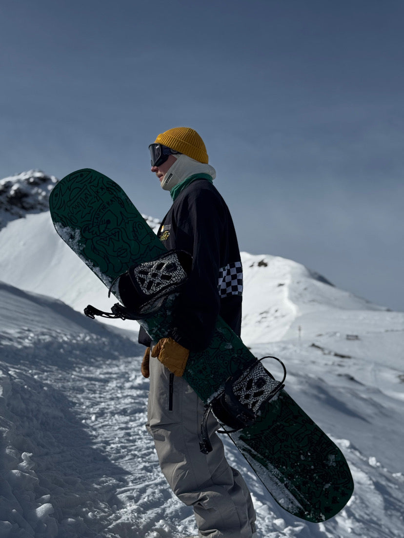 Snowboardåkare i gul stickad cozynorth mössa bär sin bräda i snötäckta fjäll, en sportig vinterstil för skidåkning och snowboard.