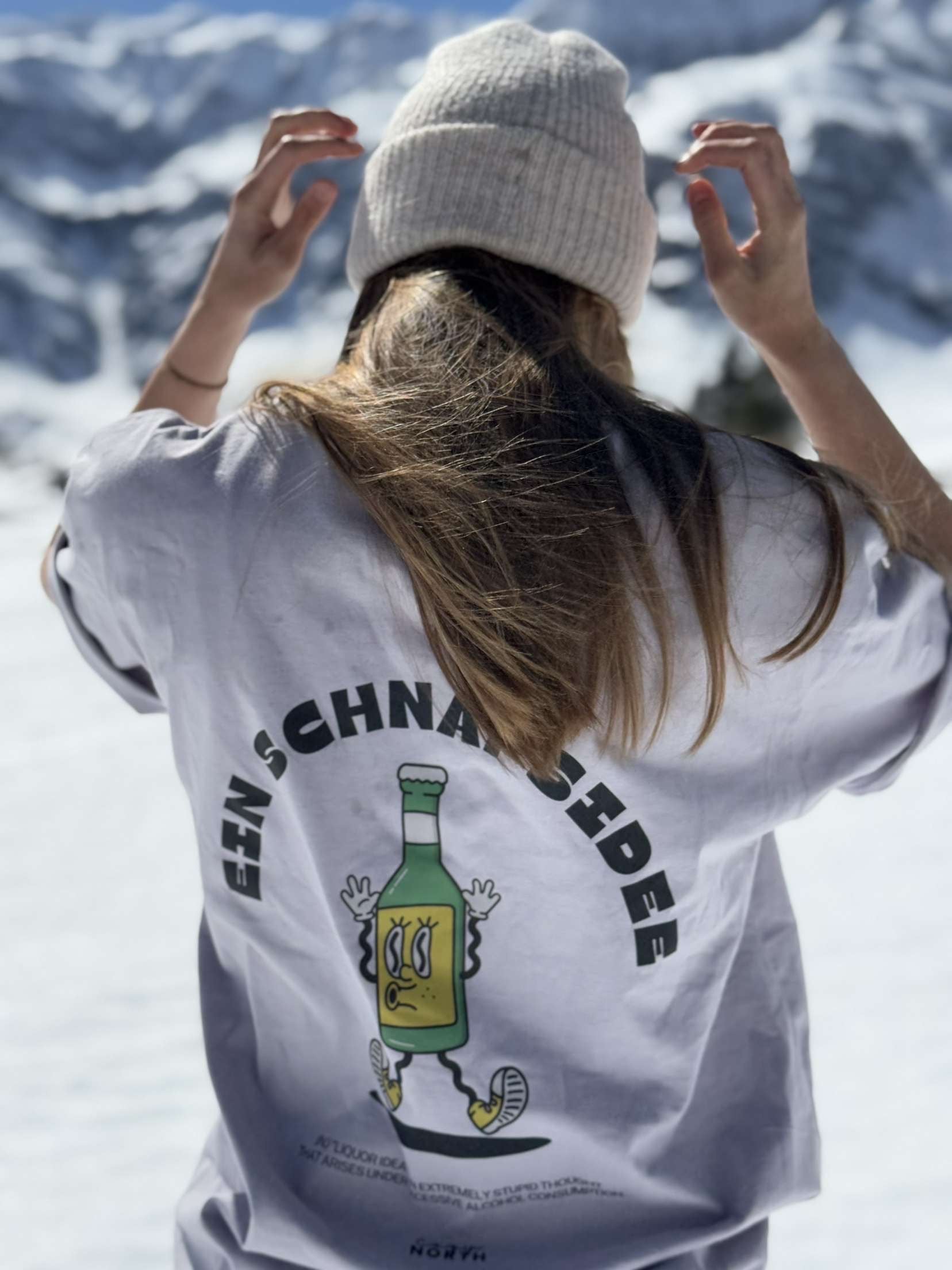 Kvinna i vit CozyNorth t-shirt med trycket ”Ein Schnapsidee” och stickad mössa, fotograferad i snöigt fjällandskap – perfekt för après-ski och vintermode.