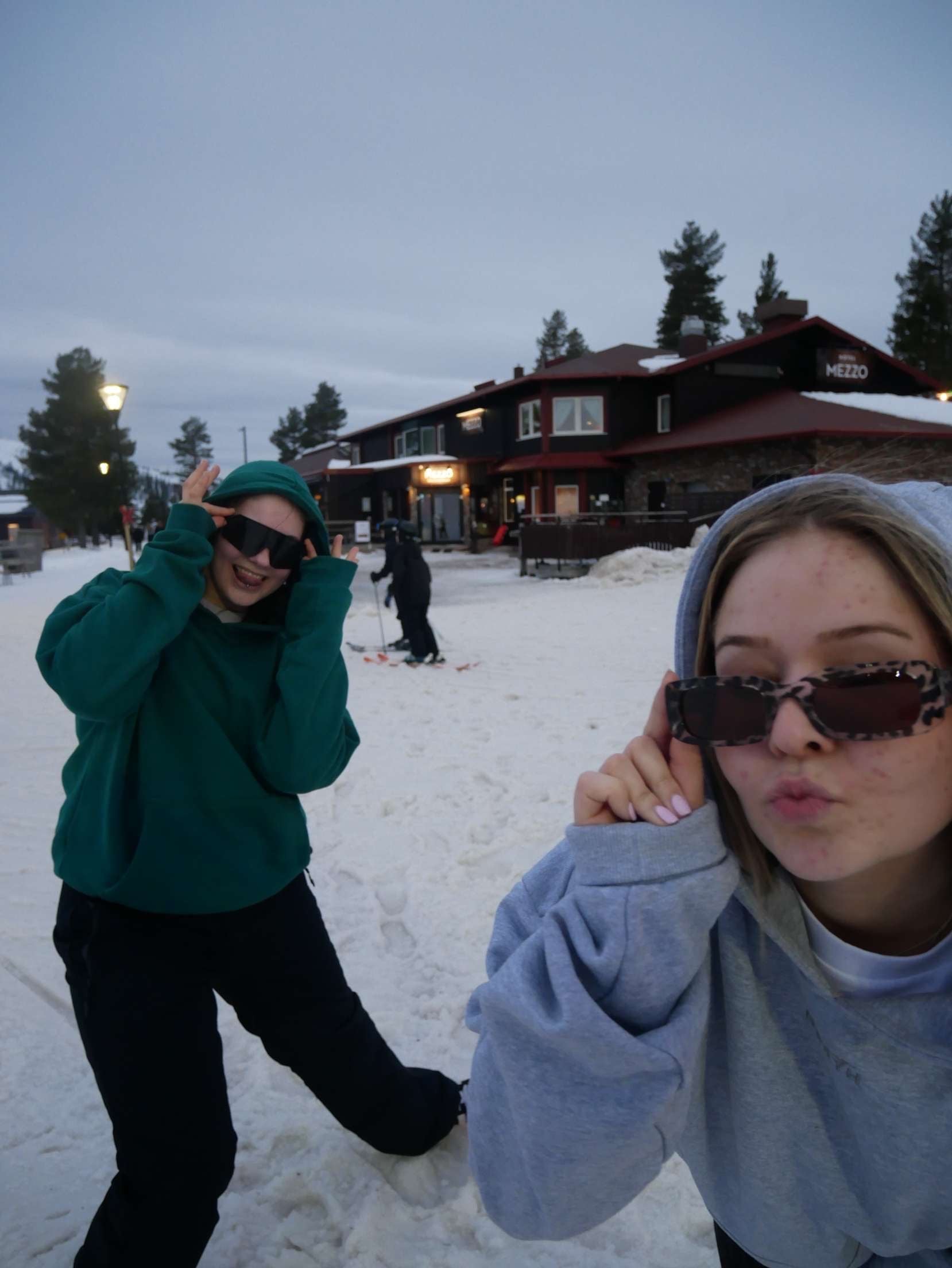 Två tjejer i CozyNorth hoodies, en grön och en grå, poserar i snön utanför afterski-stuga – trendigt vintermode för skidåkning och fjällsemester.