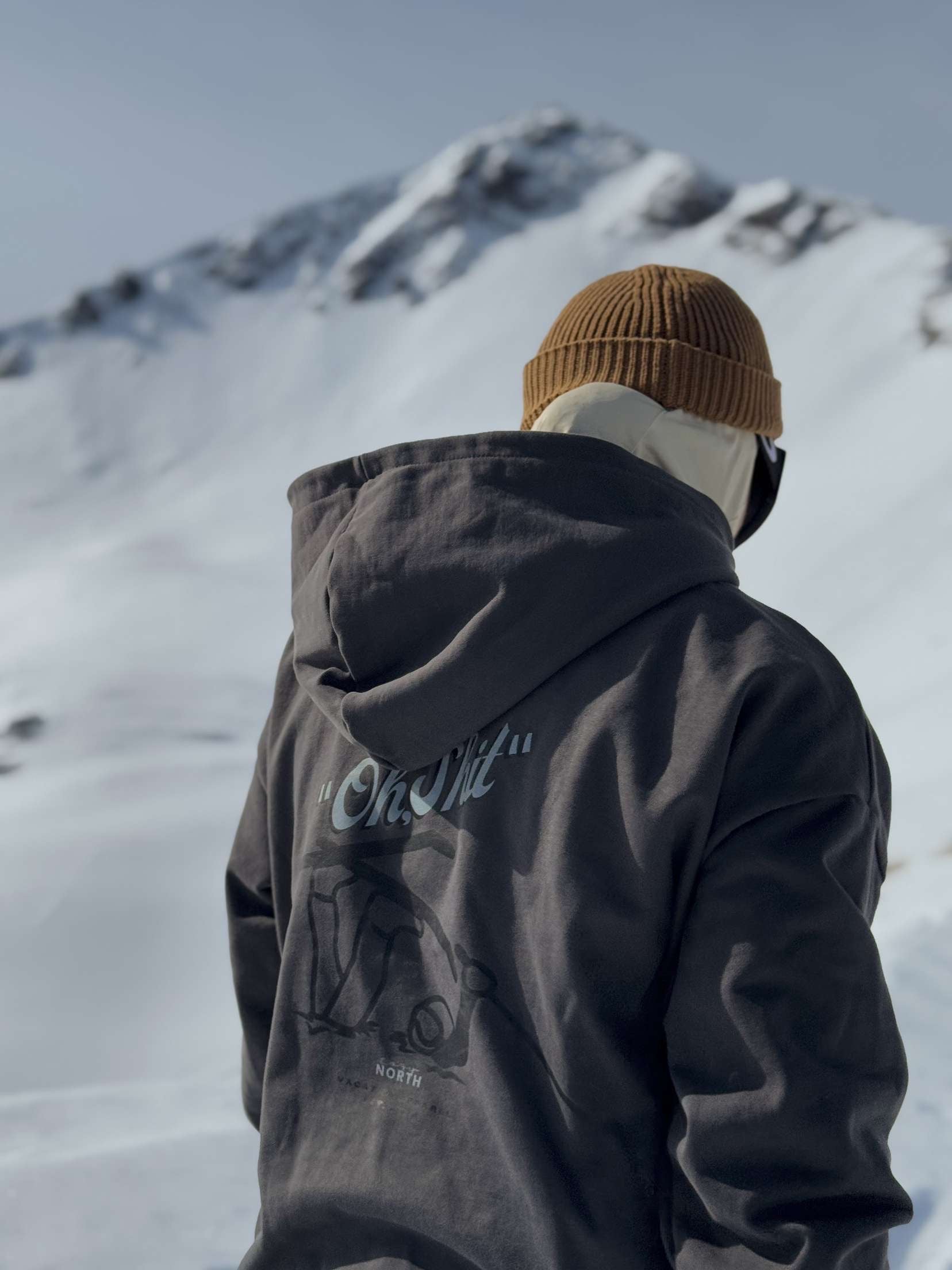 Person i svart CozyNorth hoodie med ”Oh Shit”-tryck och beige stickad CozyNorth mössa står i snötäckt fjällmiljö – en trendig vinteroutfit för skidåkning och après-ski.