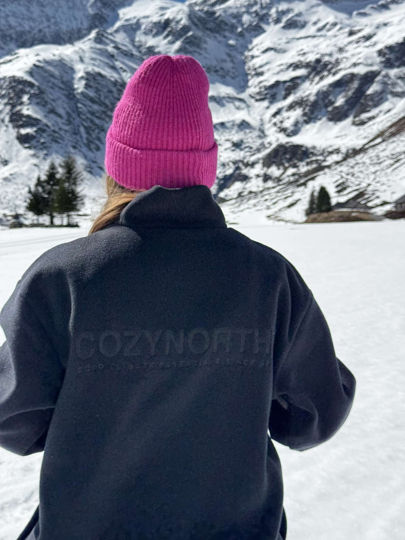 Person i svart CozyNorth fleecejacka och rosa stickad mössa står i snötäckta fjäll, en stilren vinterlook för skidåkning och après-ski.
