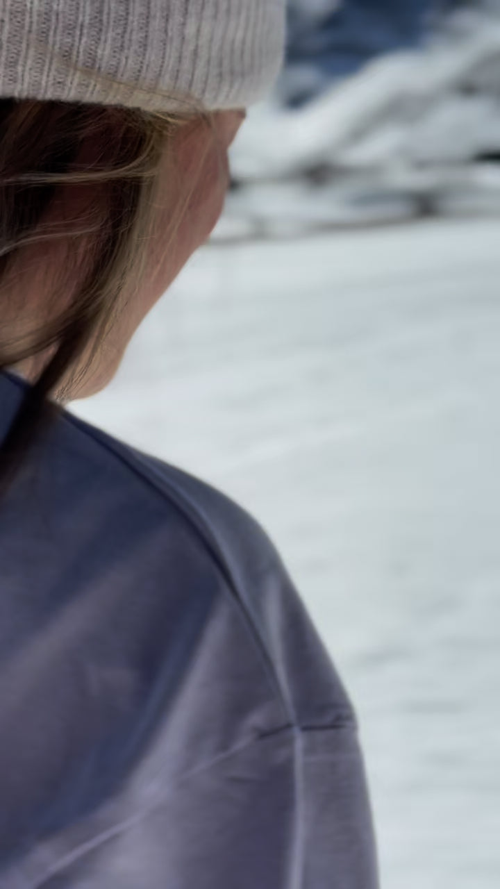 Person med vit stickad mössa och CozyNorth-tröja står i snöig fjällmiljö, en inspirerande vintervideo för skidåkning och après-ski.